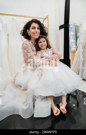 Eine freudige Braut aus dem Nahen Osten in blumigem Hochzeitskleid, die ein fröhliches Mädchen in süßer Kleidung umarmt, im Brautsalon, beim Einkaufen, bei einem besonderen Moment, Mutter und Tochter, Stockfoto