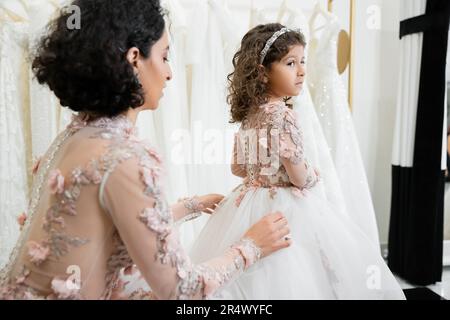 Nahöstliche Braut mit braunem, welligem Haar in geblümtem Hochzeitskleid, die den Tullrock der Tochter in süßer Kleidung anpasst, im Brautsalon, beim Einkaufen, beim Speci Stockfoto