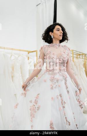 Eine bezaubernde Frau aus dem Nahen Osten, die ein wunderschönes und blumiges Hochzeitskleid anprobiert, in einem luxuriösen Brautsalon, beim Einkaufen, der zukünftigen Braut, dem verschwommenen Weiß Stockfoto
