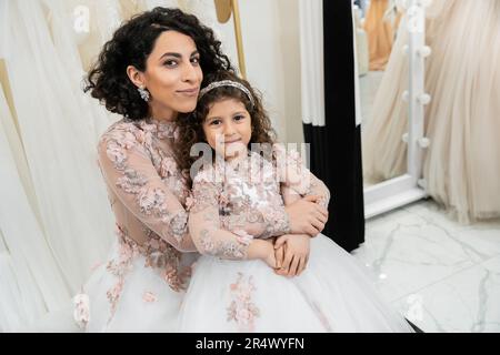Eine reizende Frau aus dem Nahen Osten in floralem Hochzeitskleid, die ein glückliches Mädchen in süßer Kleidung umarmt, im Brautsalon, beim Einkaufen, bei einem besonderen Moment, Mutter und Tochter Stockfoto