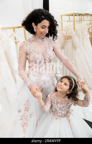 Eine reizende Braut aus dem Nahen Osten in geblümtem Hochzeitskleid, die Hand mit einem glücklichen Mädchen in süßer Kleidung hält, im Brautsalon, zum Einkaufen, besonderer Moment, Mutter Stockfoto