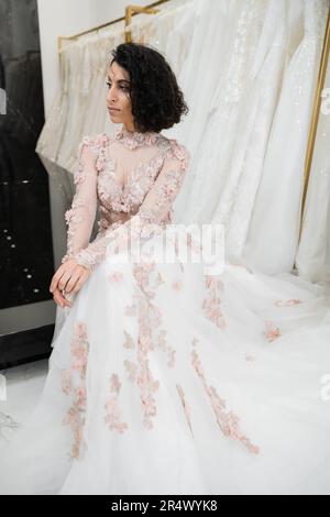 Braune Frau aus dem Nahen Osten mit welligen Haaren, die in einem wunderschönen und blumigen Hochzeitskleid neben einem verschwommenen und weißen Kleid im luxuriösen Brautsalon sitzt Stockfoto