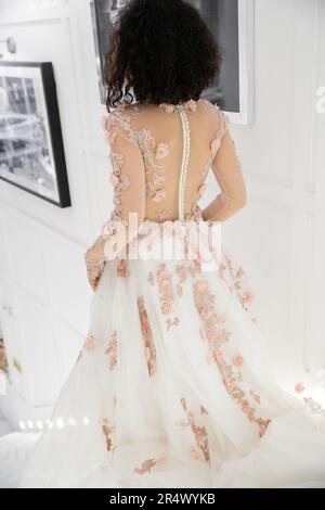 Rückansicht einer brünetten Frau aus dem Nahen Osten mit welligen Haaren, die in einem blumigen Hochzeitskleid im luxuriösen Brautssalon spaziert, eine charmante und elegante Braut Stockfoto