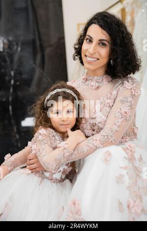 Brünette Frau aus dem Nahen Osten mit welligen Haaren, die ein süßes Mädchen umarmt und in der Nähe weißer Hochzeitskleider lächelt, mit Blumen, Mutter und Tochter, ha Stockfoto