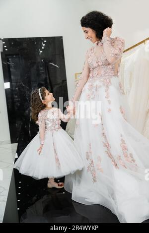 Eine fröhliche Frau aus dem Nahen Osten mit welligen Haaren in einem atemberaubenden Hochzeitskleid, die mit ihrer Tochter in süßer Blumenkleidung Händchen hält, während sie im brautsalo steht Stockfoto
