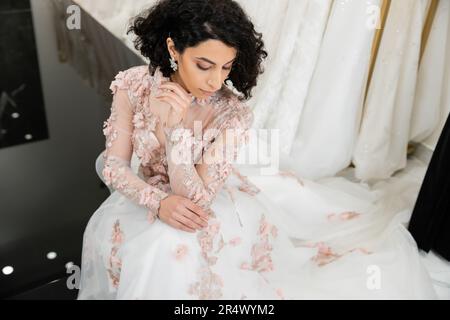 Blick von oben auf eine brünette Frau aus dem Nahen Osten mit welligen Haaren, die in einem wunderschönen und blumigen Hochzeitskleid neben einem verschwommenen und weißen Kleid im Innern des Luxus sitzt Stockfoto