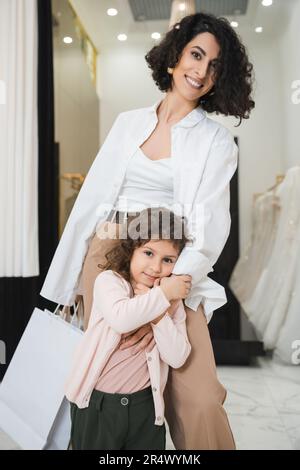 Süßes kleines Mädchen, das eine glückliche Frau aus dem Nahen Osten umarmt, mit braunem Haar und Einkaufstaschen in der Hand hält, während es in Brautkleidern neben dem Hochzeitsanzug steht Stockfoto