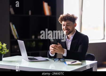 Seitenansicht eines lächelnden Geschäftsmannes, der im Büro telefoniert. Er sitzt am Desktop mit einem Laptop vor sich und tippt mit j auf dem Bildschirm Stockfoto