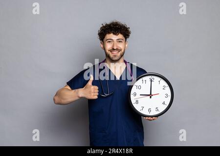 Arzt mit Daumen nach oben in der Hand halten Uhr isoliert auf einfarbigem grauen Hintergrund. Gesundheitsmedizinische Konzepte Stockfoto