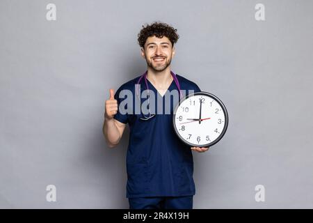 Arzt mit Daumen nach oben in der Hand halten Uhr isoliert auf einfarbigem grauen Hintergrund. Gesundheitsmedizinische Konzepte Stockfoto