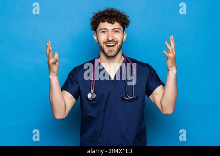 Junge gut aussehende Arzt Mann mit Bart tragen Mantel und Brille auf blauem Hintergrund feiert verrückt und verrückt für den Erfolg mit den Armen angehoben und geschlossen e Stockfoto