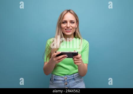 Ein bezauberndes blondes Mädchen in legerem Outfit spielt am Telefon auf blauem Hintergrund Stockfoto