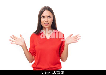 Junge weiße Frau mit schwarzem, geradem Haar in einem roten T-Shirt sieht auf weißem Hintergrund mit Kopierbereich überrascht aus Stockfoto