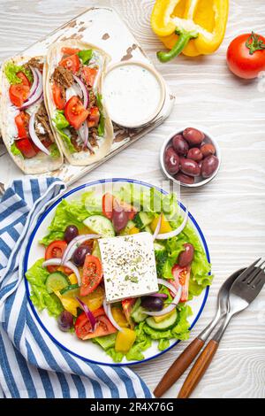 Traditionelle griechische Speisen: Griechischer Salat, Gyros mit Fleisch und Gemüse, Tzatziki-Sauce, Oliven auf weißem rustikalem Holztisch von oben Stockfoto