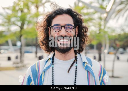 Glücklicher junger Hipster, der lächelt und in die Kamera schaut. Nahaufnahme eines individuellen Porträts von Hippie-Typ, der lacht. Vorderansicht eines bärtigen Mannes im Freien mit coolem Gesichtsausdruck. Hochwertiges Foto Stockfoto