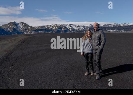 Nahaufnahme eines lächelnden Paares, das für die Kamera posiert, vor einer riesigen Landschaft aus schwarzem Sand mit einem Bergkamm und einer blauen Skipiste steht... Stockfoto