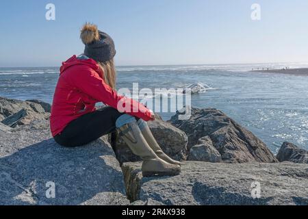 Blick auf eine Frau, die auf den Felsen am Strand sitzt und die Eisberge an der Südküste Islands vorbeiziehen sieht; Jokulsarlon, Süd-Island, Island Stockfoto