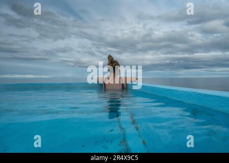 Blick von hinten auf eine Frau, die in einem heißen Pool an der Nordküste Islands sitzt und den Atlantischen Ozean überblickt Stockfoto