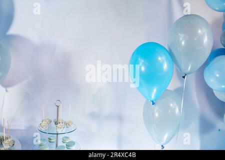 Festlicher Hintergrund mit blauen und transparenten Luftballons, Desserts, Makronen, Kuchenwürfel auf Glasschale auf hellblauem Tuch, Mockup. Thematische Geburtstagsparty Stockfoto