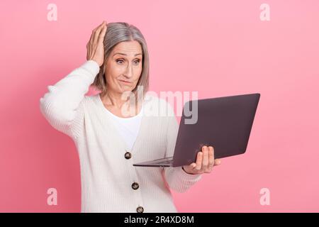 Foto in voller Länge von einer reizenden pensionierten Dame, verwirrtes, enttäuschtes Netbook in elegantem weißen Kleidungsstück, isoliert auf pinkfarbenem Hintergrund Stockfoto
