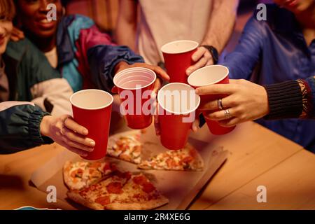 Nahaufnahme von Freunden, die rote Tassen halten und auf dem Tisch toasten, mit Pizza auf der Party, Kopierraum Stockfoto