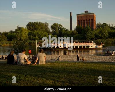 Stadtstrand in der Nähe des Flusses Odra mit vorbeifahrenden Flussbooten und Menschen, die auf dem Gras sitzen, entspannend, Breslau, Polen © Renzo Frontoni / Axiom Stockfoto