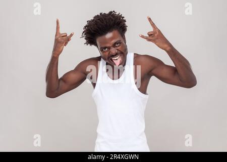Ein Mann mit Afro-Frisur schreit aus dem Glück, hat den Mund weit geöffnet, macht Rock'n Roll-Geste, sagt Rock'n'Roll diese Party. Ich trage ein weißes T-Shirt. Studioaufnahmen im Innenbereich isoliert auf grauem Hintergrund. Stockfoto
