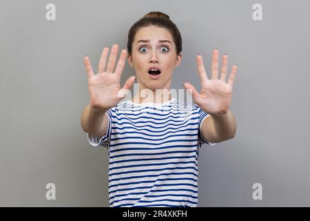 Porträt einer verängstigten jungen erwachsenen Frau, die gestreiftes T-Shirt trägt, mit Handflächen stoppt und mit verängstigten Augen überrascht aussieht. Studioaufnahmen im Innenbereich isoliert auf grauem Hintergrund. Stockfoto