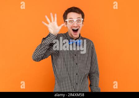 Portrait eines lustigen, aufgeregten Mannes, der seine Hand winkt, Gesten zeigt, ein Hemd mit blauer Fliege und einer weißen Brille trägt. Innenstudio, isoliert auf orangefarbenem Hintergrund. Stockfoto