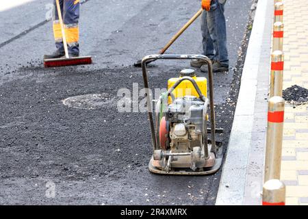 Eine alte, vibrierende Verdichtungsplatte steht am Bordstein vor dem Hintergrund einer Arbeiterbrigade von Straßenarbeitern. Stockfoto