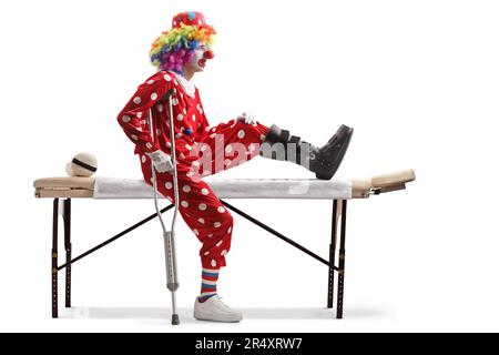 Verletzter Clown mit einer Gehstrebe auf einem Therapiebett isoliert auf weißem Hintergrund Stockfoto