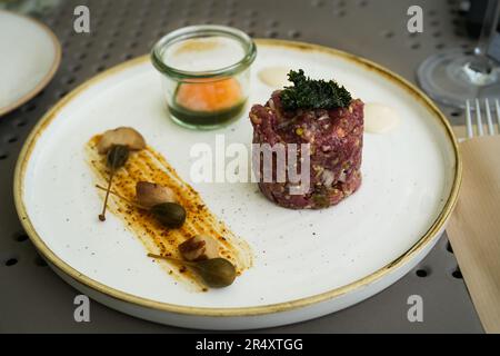 Steak Tartare mit rohem Rinderhack, Grünkohl, rohem Eigelb und Kapern auf einem weißen Teller mit goldenem Rand. Keto Diät, roh gesunde Essen Gourmet Stockfoto