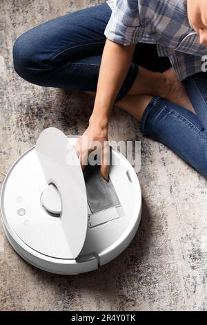 Das Mädchen nimmt den Staubbehälter aus dem Staubsaugerroboter, um ihn zu reinigen. Das Konzept eines intelligenten Hauses, Hausarbeit, Reinigung, Wohnung behalten Stockfoto