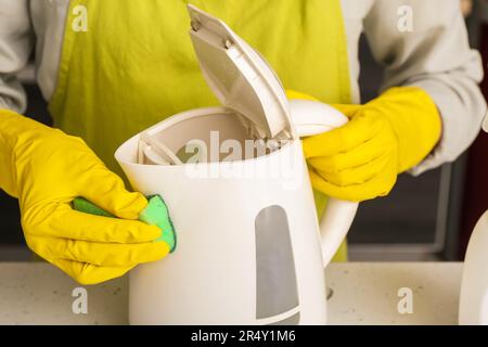 Frau in gelben Handschuhen, die elektrischen Wasserkocher auf einem weißen Tisch reinigt Stockfoto