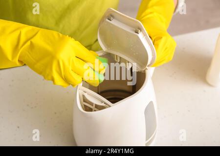 Frau in gelben Handschuhen, die elektrischen Wasserkocher auf einem weißen Tisch reinigt Stockfoto
