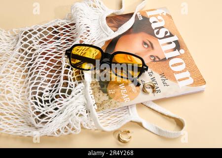 Stilvolle Sonnenbrille und Tragetasche mit Zeitschrift auf beigefarbenem Hintergrund Stockfoto