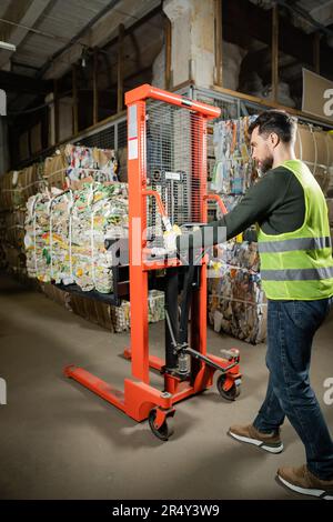 Seitenansicht des bärtigen Sortierers in Schutzweste und Handschuhen Verwendung eines Handhubwagens mit Altpapier bei der Arbeit in einer verschwommenen Müllsortierstelle, RE Stockfoto