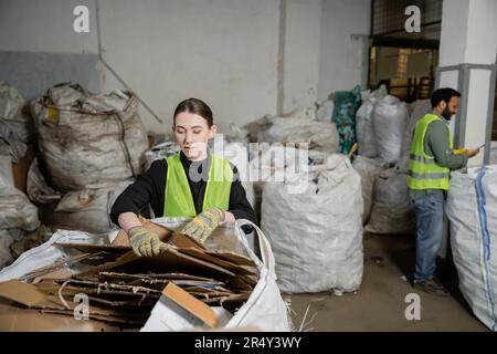 Junge Mitarbeiterin in Schutzweste und Handschuhen, die Altpapier in den Sack legt, während sie in der Nähe einer unscharfen indischen Kollegin in der Abfallentsorgungsstation, g Stockfoto