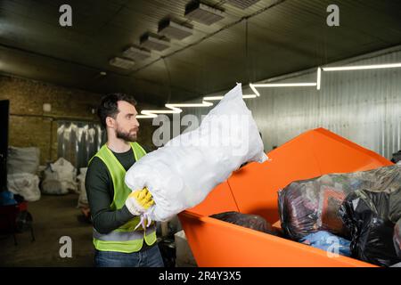 Seitenansicht eines Mannes in Warnweste und Handschuh beim Verstauen des Sacks mit Müll im Behälter während der Arbeit in der Abfallentsorgungsstation, Mülltrennung und Stockfoto