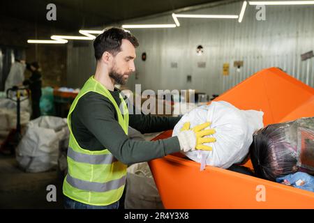 Seitenansicht des Sortierers in Handschuhen und Schutzweste Sack in Behälter legen während der Arbeit in verschwommener Abfallentsorgungsstation im Hintergrund, Müllsortierung Stockfoto