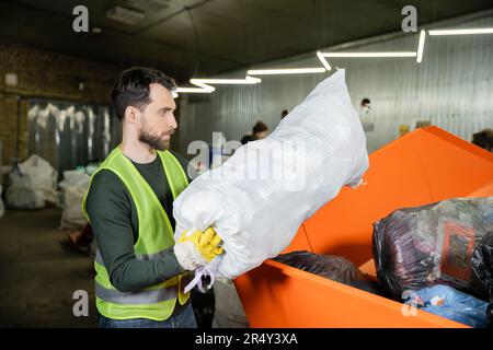 Seitenansicht des bärtigen Sortierers in Handschuhen und Schutzweste, Ablagesack mit Müll in Box in verschwommener Abfallentsorgungsstation, Müllsortierung und Recycling Stockfoto