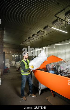 Seitenansicht des bärtigen männlichen Arbeiters in Handschuh und fluoreszierende Weste, den Sack mit Müll in Behälter in verschwommene Abfallentsorgungsstation, Müllsortierung Stockfoto