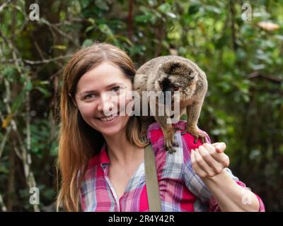 Junge Frau im Hemd, posiert mit einem kleinen Lemur auf der Schulter, lächelnd, verschwommener Dschungelbaum-Hintergrund Stockfoto