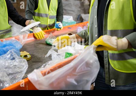 Zugeschnittene Ansicht des Sortierers in Handschuhen und Schutzweste mit verschwommenem Plastikbeutel und Entnahme von Müll vom Förderband bei der Arbeit in der Nähe von Kollegen im Abfallbereich Stockfoto