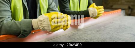 Zugeschnittene Ansicht des Sortierers in Schutzhandschuhen und einer Weste, die neben dem Förderer steht, und verschwommener Kollege bei der Arbeit in der Müllsortieranlage, Müllcontainer Stockfoto