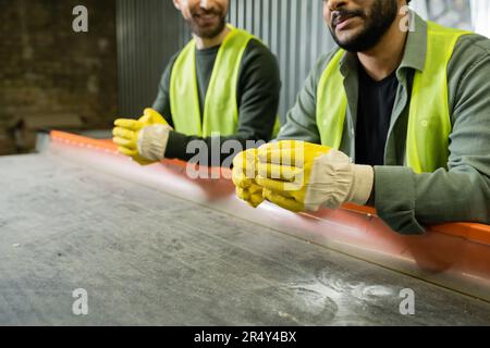 Gekürzter Blick auf den indischen Sortierer in gut sichtbarer Weste und Handschuhen, die neben dem Förderer stehen und einen verschwommenen Kollegen bei der Arbeit im Müllsortierzentrum, Stockfoto