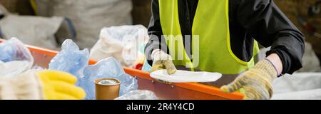 Zugeschnittene Ansicht des Sortierers in Schutzweste und Handschuhen, die Müll vom Förderband während der Arbeit in der Abfallentsorgungsstation im Hintergrund, Müllsorti Stockfoto