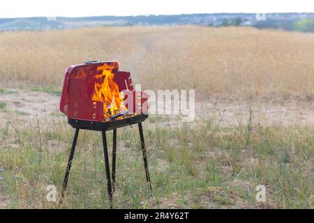 Mobiler Grill oder Barbecue auf einem Boden in der Nähe des Weizenfeldes. Sichtbare Flammen und Feuer auf einem Brennstoff aus Holz und Kohle. Stockfoto