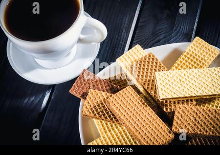 Kaffee in einer Tasse mit Keksen auf einem Teller im Hintergrund. Kaffee mit Waffeln auf einem schwarzen Holztisch. Stockfoto