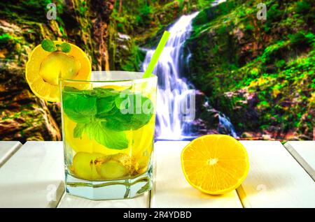 Trinken Sie ein Glas auf einem weißen Holztisch. Drink mit Zitrone, Ingwer und Minze mit Berg-Wasserfall im Hintergrund. Stockfoto